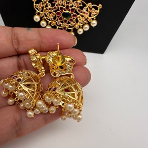May include: Gold-toned, ornate earrings with a bell shape, featuring red and clear gemstones, and dangling faux pearls. The earrings are held in a hand, with a matching pendant in the background.