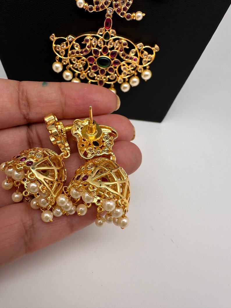May include: Gold-toned, ornate earrings with a bell shape, featuring red and clear gemstones, and dangling faux pearls. The earrings are held in a hand, with a matching pendant in the background.