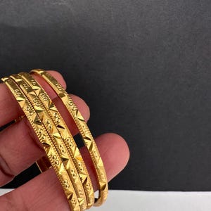 May include: Four gold bangles with detailed geometric patterns. The bangles are displayed against a black and white background, highlighting their intricate design and polished finish. A traditional piece of jewellery.