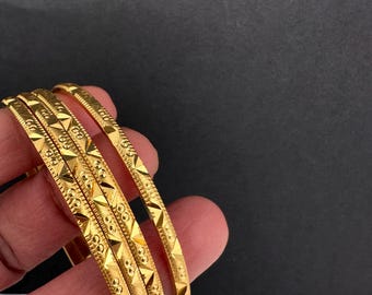 Könnte beinhalten: Ein Satz von vier goldenen Armreifen mit aufwendigen geometrischen Mustern. Die Armreifen werden vor einem schwarz-weißen Hintergrund präsentiert, um ihr detailliertes Design und ihre glänzende Oberfläche hervorzuheben. Klassischer Schmuck.