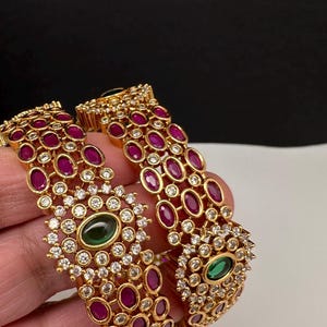 May include: Gold-toned bangles with ruby-red and clear crystal accents. Each bangle features a central green gemstone surrounded by crystals. The design includes rows of oval-shaped red stones.