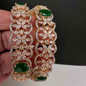 May include: Two gold-tone bangles featuring a repeating floral pattern set with clear marquise-cut stones and accented with oval-shaped green stones. The metal framework is encrusted with small clear stones along the edges of the floral motifs.