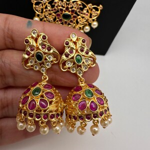 May include: Gold-toned, ornate earrings with ruby-red and emerald-green accents. The earrings feature a bell-shaped design with dangling faux pearls. A matching pendant is visible in the background, showcasing similar detailing.