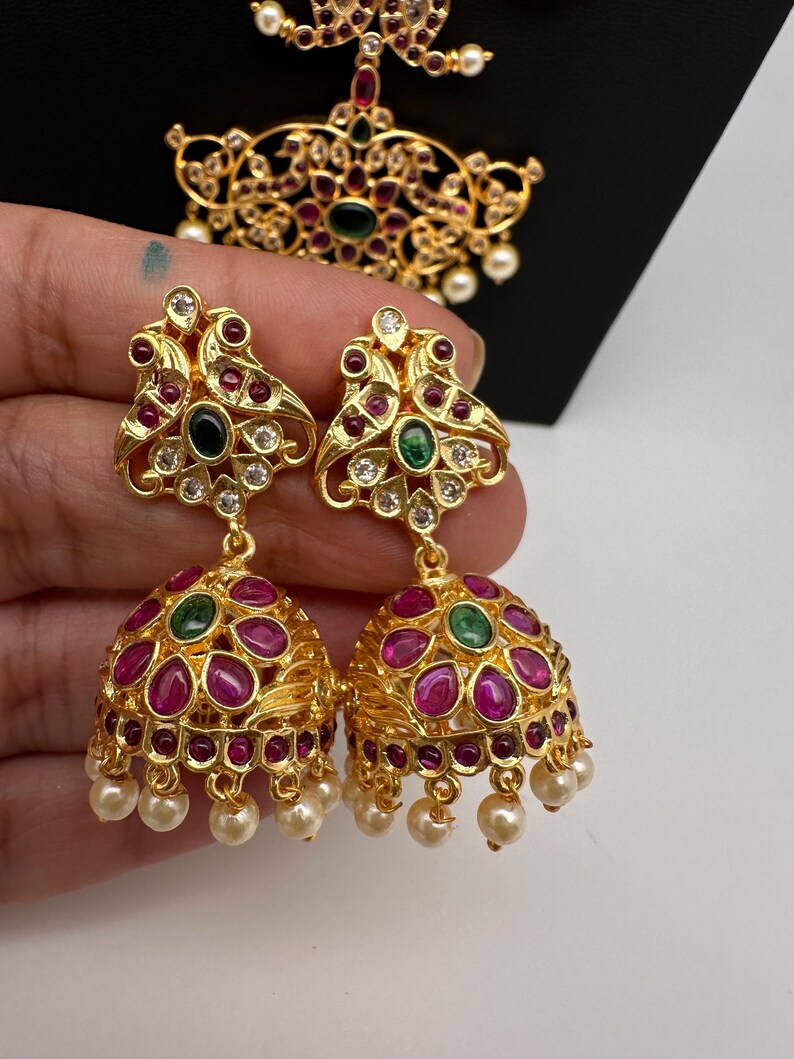 May include: Gold-toned, ornate earrings with ruby-red and emerald-green accents. The earrings feature a bell-shaped design with dangling faux pearls. A matching pendant is visible in the background, showcasing similar detailing.