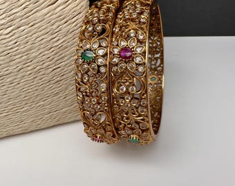 Puede incluir: Dos pulseras doradas con intrincados diseños florales. Cada pulsera presenta piedras preciosas incrustadas en colores verde, rojo y transparente. Las pulseras se exhiben sobre una superficie blanca, mostrando su detallada artesanía y estilo ornamentado.