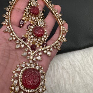 May include: Gold-coloured bridal jewellery set with a headpiece and earrings. The set features red carved stone accents, clear crystals, and faux pearls. The headpiece has a curved design with crystal embellishments. The earrings have a similar design with dangling elements.