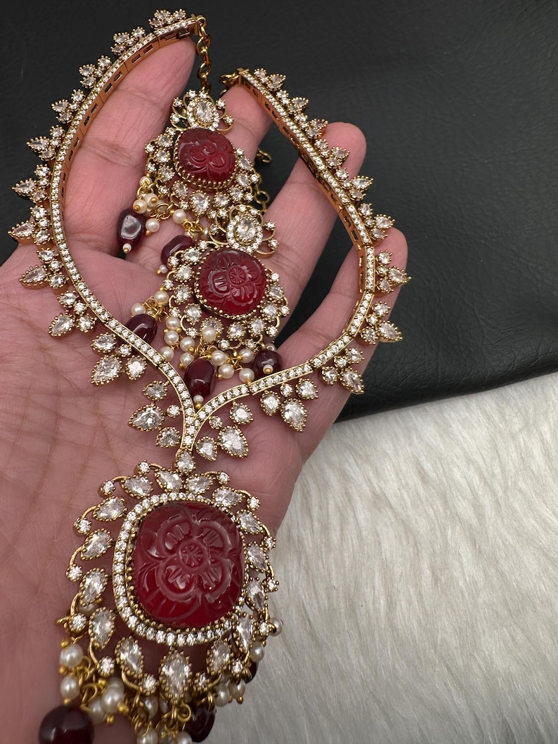 May include: Gold-coloured bridal jewellery set with a headpiece and earrings. The set features red carved stone accents, clear crystals, and faux pearls. The headpiece has a curved design with crystal embellishments. The earrings have a similar design with dangling elements.