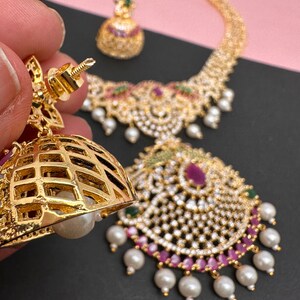 Op de afbeelding: Een goudkleurige sieradenset met een ketting, oorbellen en een hanger. De ketting en hanger zijn versierd met heldere en robijnkleurige stenen, groene accenten en bungelende witte parels. De oorbellen hebben een tralieontwerp met een parel.