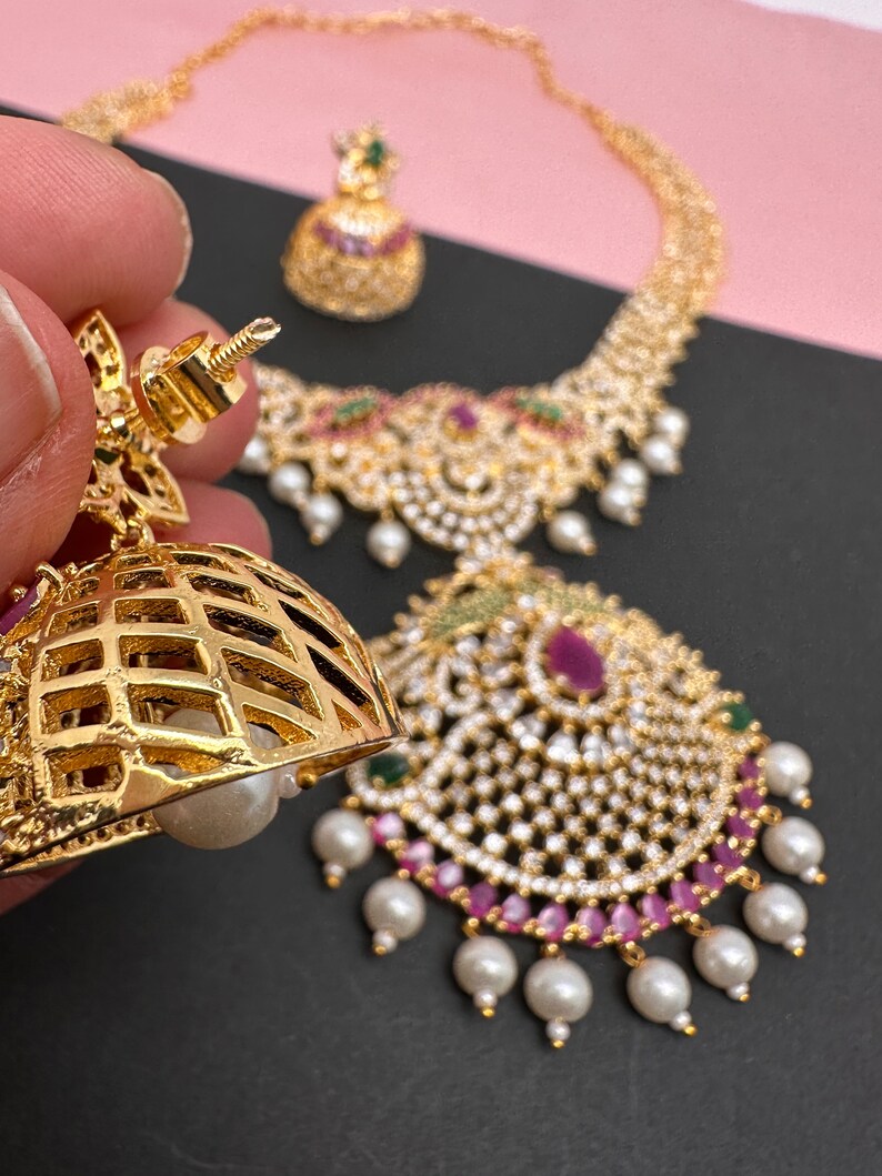 Op de afbeelding: Een goudkleurige sieradenset met een ketting, oorbellen en een hanger. De ketting en hanger zijn versierd met heldere en robijnkleurige stenen, groene accenten en bungelende witte parels. De oorbellen hebben een tralieontwerp met een parel.