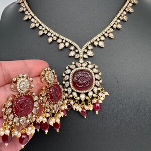 May include: A gold-coloured necklace and earring set. The necklace has a pendant with a carved red stone surrounded by clear crystals. The earrings have matching red stones and crystal accents, with dangling red beads and small white pearls.