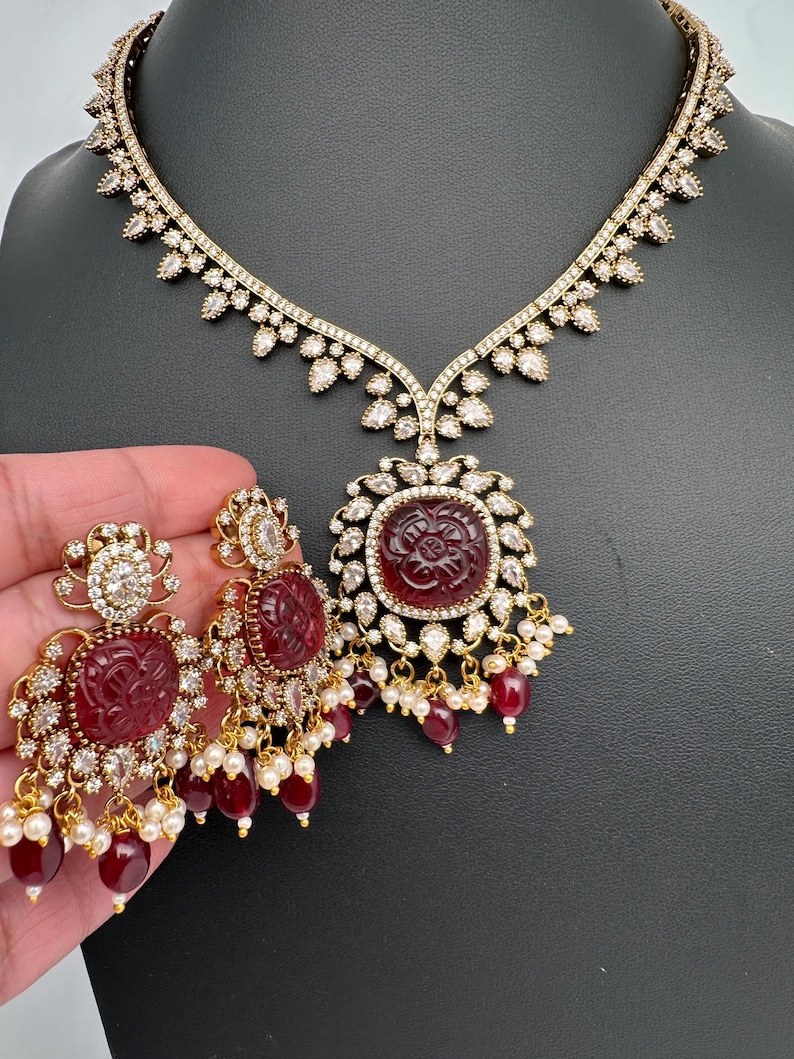 May include: A gold-coloured necklace and earring set. The necklace has a pendant with a carved red stone surrounded by clear crystals. The earrings have matching red stones and crystal accents, with dangling red beads and small white pearls.