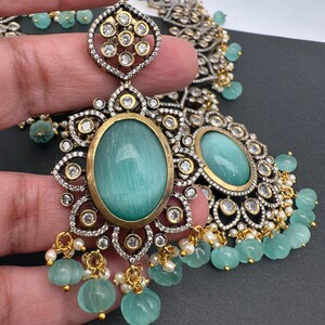 May include: An ornate silver and gold-toned jewelry set featuring large oval, light blue-green gemstones. The earrings and necklace have intricate detailing with clear, round accent stones and small, dangling, carved green beads.