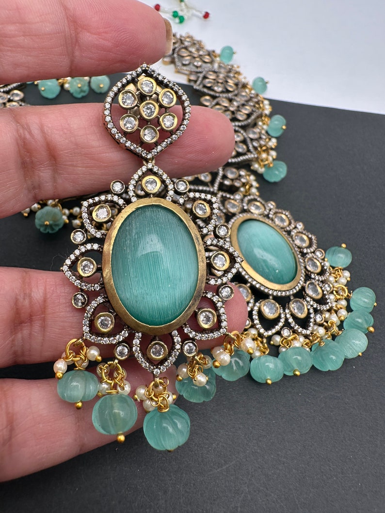 May include: An ornate silver and gold-toned jewelry set featuring large oval, light blue-green gemstones. The earrings and necklace have intricate detailing with clear, round accent stones and small, dangling, carved green beads.