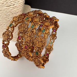 May include: Ornate gold-colored bangles with intricate designs and embedded gemstones. The bangles feature detailed patterns and are adorned with red and green gemstones. The jewelry is displayed on a white surface.