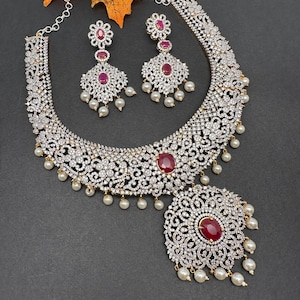 May include: A dazzling jewelry set featuring a necklace and matching earrings. The necklace and earrings are adorned with sparkling clear stones, ruby-red accents, and dangling faux pearls. The necklace has a large, ornate pendant. The set is displayed on a dark background.