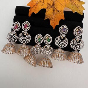 May include: A collection of ornate, gold-toned and silver-toned jhumka earrings. Each earring features a bell-shaped bottom and a detailed, filigree design with embedded clear and colored gemstones. A large, yellow leaf rests above the earrings.