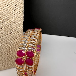 May include: Two gold bangles, each featuring alternating rows of clear crystals and large, oval ruby-red stones. The bangles are displayed against a neutral background with a woven texture on the left.