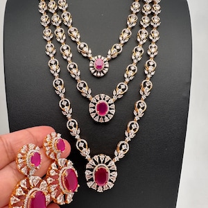 May include: A three-piece jewelry set featuring a necklace and earrings. The necklace has three strands with a floral design, and the earrings have a matching design. The jewelry is gold-toned with clear and ruby-colored stones.