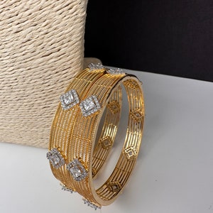 May include: Gold bangles with an openwork design. Each bangle has square, clear gemstones and diamond-shaped accents. The jewellery is displayed on a white surface with a textured, beige background.