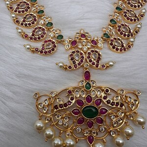 May include: A gold-toned necklace and pendant set. The necklace features paisley-shaped designs with red and clear gemstones, green accents, and small pearls. The pendant has a large green center stone, red and clear gemstones, and dangling pearls.