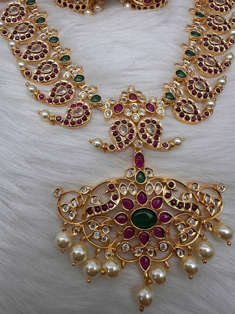 May include: A gold-toned necklace and pendant set. The necklace features paisley-shaped designs with red and clear gemstones, green accents, and small pearls. The pendant has a large green center stone, red and clear gemstones, and dangling pearls.
