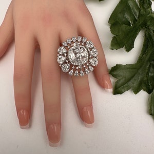 May include: A large, silver-toned ring with a central square-cut clear stone, surrounded by smaller, round and pear-shaped clear stones. The ring is set on a light-toned surface.