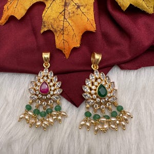 May include: Two gold-toned pendant earrings. Each features a central gemstone, one red and one green, surrounded by clear crystals. Green beads and small pearl-like drops dangle below. The earrings are set against a white, fluffy background.
