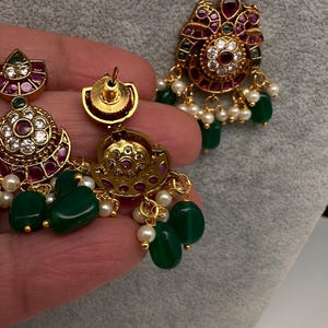 May include: Gold-toned jewelry set featuring a necklace and earrings. The pieces are adorned with green, red, and white gemstones, and small white pearls. The earrings and pendant have a floral design.