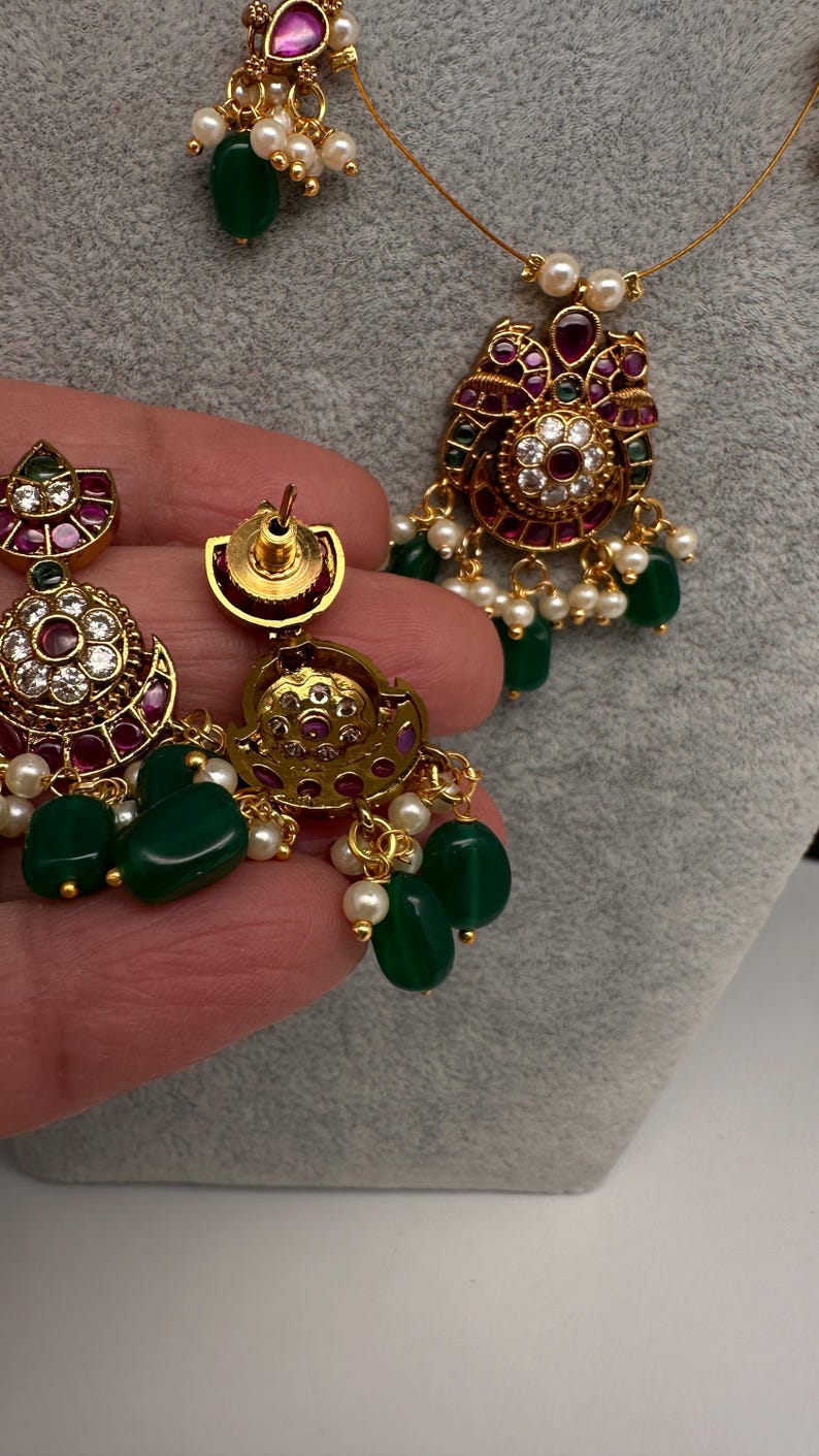 May include: Gold-toned jewelry set featuring a necklace and earrings. The pieces are adorned with green, red, and white gemstones, and small white pearls. The earrings and pendant have a floral design.
