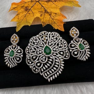 May include: A jewelry set featuring a pendant and matching earrings. The pendant and earrings are adorned with clear, sparkling stones and a central green gemstone. The set is displayed on a black surface with a decorative yellow leaf.
