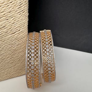 May include: Two gold-toned bangles with intricate openwork and embedded clear gemstones. The bangles have a two-tone design, with gold and silver-colored metal. The jewelry is displayed on a white surface with a woven texture in the background.