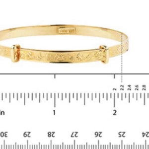Puede incluir: Una pulsera tipo brazalete extensible de tono dorado con una serie de peque&ntilde;os s&iacute;mbolos grabados a lo largo de la banda. La pulsera est&aacute; colocada sobre una regla, mostrando un di&aacute;metro de aproximadamente 5,6 cm en la secci&oacute;n central.