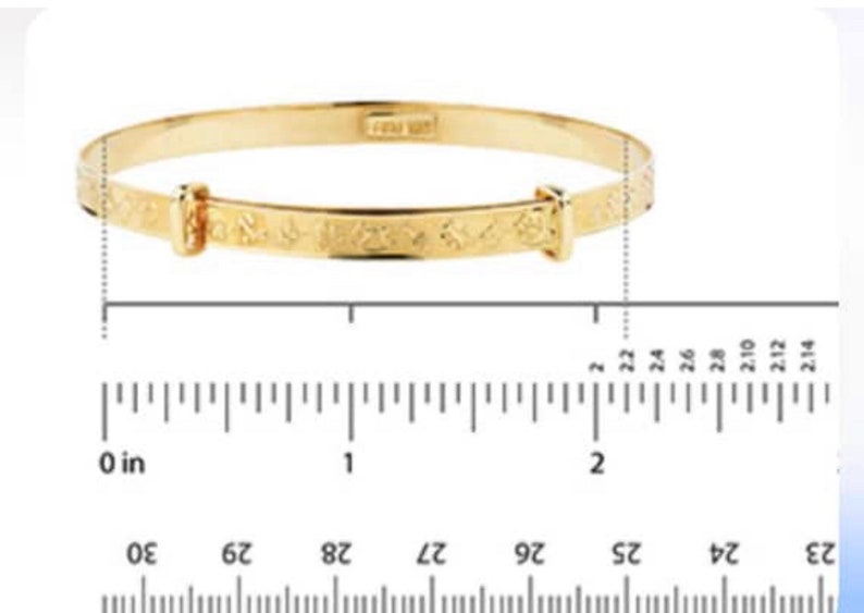 Puede incluir: Una pulsera tipo brazalete extensible de tono dorado con una serie de peque&ntilde;os s&iacute;mbolos grabados a lo largo de la banda. La pulsera est&aacute; colocada sobre una regla, mostrando un di&aacute;metro de aproximadamente 5,6 cm en la secci&oacute;n central.