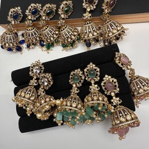 May include: An assortment of gold-toned, ornate earrings with various gemstone accents. The earrings feature intricate detailing, including clear crystals and dangling beads. Gemstones include blue, green, purple, and pink. The earrings are displayed on a black surface.