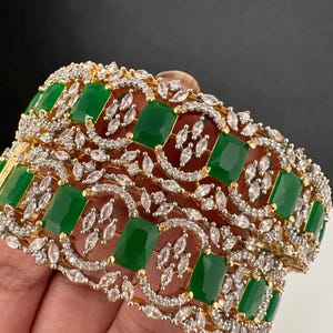 May include: Two gold-toned bangles with emerald green rectangular stones and clear, sparkling accents. The design features a repeating pattern of green stones and clear crystals, creating a luxurious look. The bangles are held against a dark background.