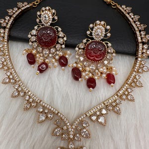 May include: A gold-coloured jewellery set comprising a necklace and earrings. The necklace and earrings feature a floral design with red gemstone centrepieces, surrounded by clear crystals and small pearl accents. The set is displayed on a white, textured surface.