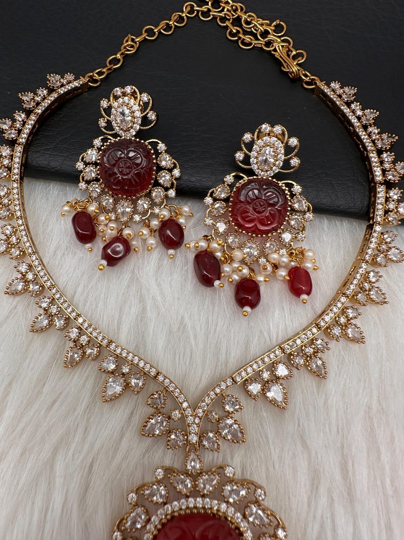 May include: A gold-coloured jewellery set comprising a necklace and earrings. The necklace and earrings feature a floral design with red gemstone centrepieces, surrounded by clear crystals and small pearl accents. The set is displayed on a white, textured surface.