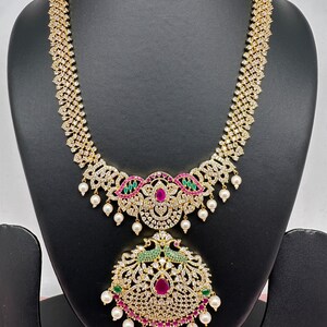 Op de afbeelding: Een goudkleurige ketting met een hanger met een pauwontwerp. De ketting is versierd met heldere kristallen, rode en groene edelstenen en bungelende witte parels. De ketting wordt getoond op een zwarte mannequin.