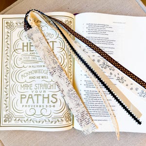 Peut inclure: Une Bible ouverte avec le texte de Proverbes 3:5-6 illustré sur la page de gauche et du texte sur la droite. Cinq marque-pages en ruban décoratif aux motifs variés, incluant du texte, des pois et des fleurs, sont fixés à la reliure.
