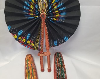 May include: A circular hand fan with a black fabric base featuring a vibrant yellow, blue, and red patterned design. The fan has a brown leather handle and frame. Two additional folded fans with matching leather handles are positioned in the foreground.