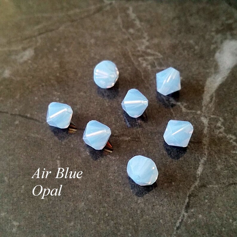 May include: Seven light blue iridescent glass beads with a faceted design. The beads are arranged in a semi-circle on a gray surface. The text "Air Blue Opal" is written below the beads.