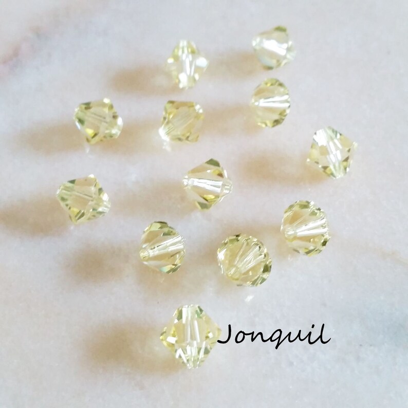 May include: A close-up of 12 small, clear, faceted glass beads in a pale yellow color. The beads are arranged in a scattered pattern on a white surface. The word "Jonquil" is written in black lettering below the beads.