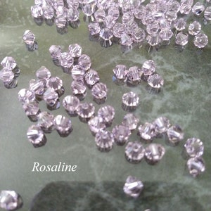 May include: A large quantity of small, clear, faceted glass beads in a light pink color. The beads are scattered on a reflective surface. The word "Rosaline" is written in white text on the surface.