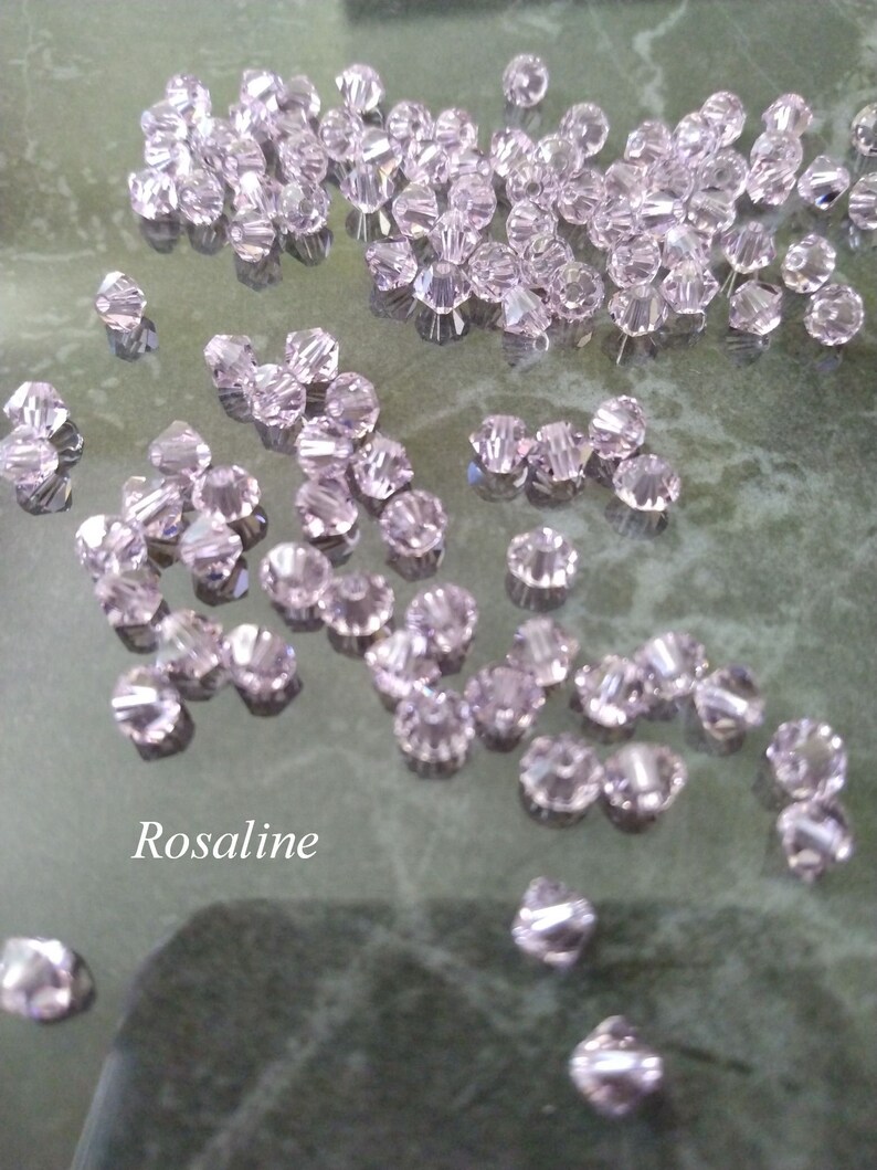 May include: A large quantity of small, clear, faceted glass beads in a light pink color. The beads are scattered on a reflective surface. The word "Rosaline" is written in white text on the surface.