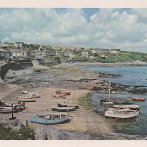 May include: A scenic coastal view featuring a sandy beach with several small boats resting on the shore. Buildings line the coast, and a green hillside rises in the background under a cloudy sky. The water is a clear turquoise color.