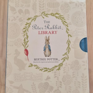 Puede incluir: Un estuche de libros de "The Peter Rabbit Library". La portada presenta una corona verde que rodea el título y una ilustración de Peter Rabbit. El fondo es de color beige claro con ilustraciones florales y de animales.