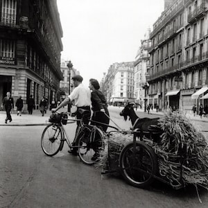 May include: Black and white photograph of a street scene featuring a bicycle pulling a cart loaded with hay, and a goat. A man and a woman are walking alongside the bicycle. Buildings line the street.