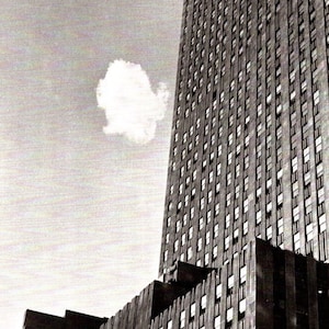 Puede incluir: Fotografía en blanco y negro de un rascacielos con numerosas ventanas. La fachada del edificio es texturizada y una nube flota en el cielo. La parte inferior del edificio tiene un diseño escalonado.