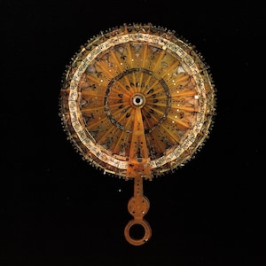 May include: A detailed, antique fan with a circular design. The fan features a golden-yellow color with intricate patterns and embellishments. The handle has a loop at the end. The background is black.