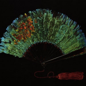 May include: A decorative hand fan with a dark wooden handle and a green and blue feathered design. The fan features a floral pattern in shades of orange and red. A red tassel hangs from the base of the fan against a black background.